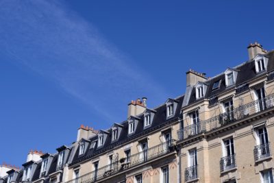 Une rangée d'immeubles d'appartements parisiens traditionnels aux toits d'ardoise et aux multiples cheminées sous un ciel bleu clair, telle que photographiée par le célèbre photographe Thomas Duclert. Les bâtiments présentent des balcons en fer forgé et une architecture classique.
