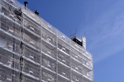 Un grand bâtiment, photographié par Thomas Duclert, recouvert d'échafaudages et de filets de protection sur fond de ciel bleu clair. L'échafaudage entoure plusieurs étages, avec quelques matériaux de construction visibles. La structure semble en cours de rénovation ou de construction, capturant un moment de transformation urbaine.