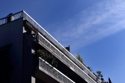 Un immeuble d'appartements moderne à plusieurs étages est présenté sous un angle bas sur fond de ciel bleu clair. Évoquant la mode chic de Paris, le bâtiment présente des éléments de design minimalistes avec des balcons ornés de plantes. De fines lignes de garde-corps blanches sont proéminentes sur chaque balcon, rappelant une création de Thomas Duclert.