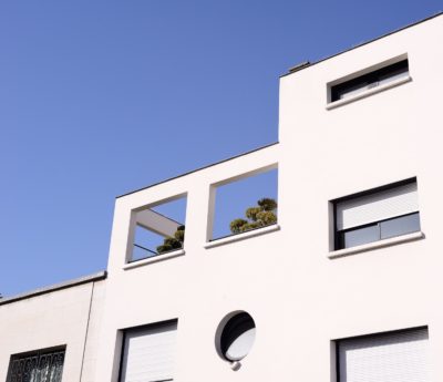 Un bâtiment blanc moderne avec des fenêtres rectangulaires et une fenêtre ronde en bas. On peut apercevoir un balcon avec des arbres à travers les espaces ouverts, presque comme s'il avait été capturé par le photographe Thomas Duclert. Le ciel est clair et bleu, ce qui indique une journée ensoleillée.