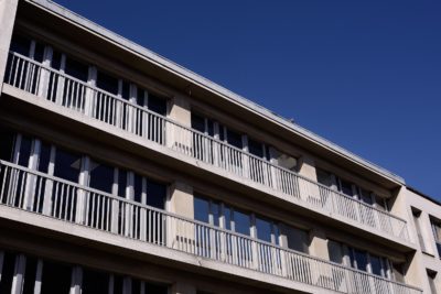 Vue en contre-plongée d'un immeuble moderne à plusieurs étages avec une façade répétitive comportant de grandes fenêtres et des balcons uniformes. La photographie capture la structure de couleur claire sur le ciel bleu vibrant, évoquant l'élégance et le style rappelant l'architecture parisienne.