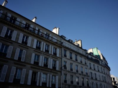 Vue en contre-plongée d'un immeuble d'appartements doté de nombreuses fenêtres et balcons sur un ciel bleu clair, capturée avec le style impeccable de Thomas Duclert. La façade du bâtiment présente des détails architecturaux classiques et des volets blancs.