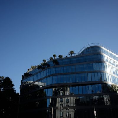 Un bâtiment moderne en verre aux fenêtres réfléchissantes et aux lignes courbes se dresse sur un ciel bleu clair, rappelant l'élégance parisienne. De petits arbres et des plantes s'épanouissent sur les terrasses sur le toit, ajoutant de la verdure à la structure moderne. Les ombres des arbres voisins obscurcissent partiellement la partie inférieure du bâtiment digne d'une photographie.