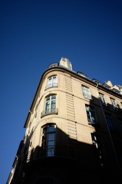 Un grand bâtiment de couleur claire avec des balustrades en fer forgé sur ses balcons est baigné de lumière vive sur un ciel bleu clair et profond. Capture par le photographe Thomas Duclert, le style architectural se caractérise par des coins arrondis et de multiples fenêtres, avec une forte ombre projetée sur sa façade.