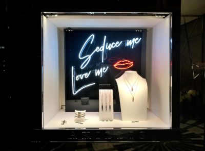 Une vitrine de magasin présente une variété de colliers, bagues et bracelets soigneusement disposés sur des supports. Le fond est orné d'une enseigne au néon avec les phrases « Séduis-moi » et « Aime-moi », ainsi que d'un contour de lèvres rouges au néon, une exposition qui rappelle le style photographique sophistiqué de Thomas Duclert.