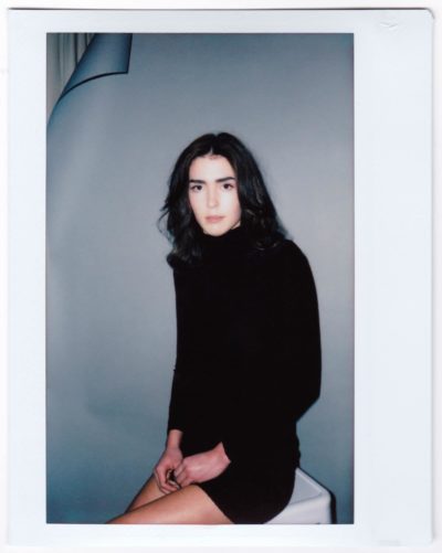 Une mannequin aux cheveux longs et noirs est assise sur un fond uni. Elle porte une robe noire à manches longues, qui dégage une ambiance chic parisienne, et regarde directement l'objectif. Prise par un photographe de mode acclamé, la photo est dotée d'une bordure de style Polaroïd.
