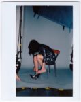 Une mannequin aux cheveux noirs est assise sur une chaise dans un studio de photographie, se penchant pour enfiler des chaussures noires à talons hauts. La mannequin, vêtue d'une robe brillante de couleur sombre, se tient debout sur un fond gris avec du matériel visible autour d'elle - une scène classique capturée par le photographe Thomas Duclert.