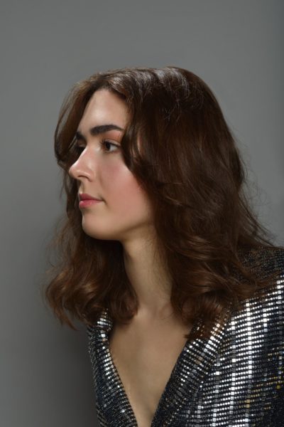 Une mannequin aux cheveux bruns ondulés, longs jusqu'aux épaules, regarde vers la gauche sur un fond gris uni. Elle porte un haut argenté à paillettes avec un décolleté profond, lui donnant une apparence glamour, telle que capturée par un photographe de mode expérimenté à Paris.
