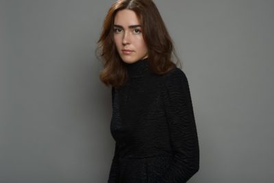 Une mannequin se tient debout sur un fond gris uni. Elle a les cheveux bruns ondulés jusqu'aux épaules et porte un haut noir à manches longues. Avec une expression sérieuse, ce modèle évoque l'essence du style parisien vu à travers l'objectif de Thomas Duclert.