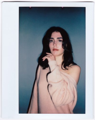 Une mannequin aux cheveux noirs mi-longs pose devant un fond neutre dans une séance photo inspirée de la mode. Elle porte un pull rose clair oversize à épaules dénudées et regarde directement l'objectif. Sa main droite effleure doucement son cou, évoquant l'élégance du style parisien.