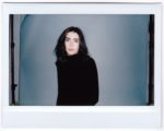 Une mannequin aux cheveux noirs mi-longs, portant un col roulé noir, se tient debout sur un fond gris. L'image, prise par le photographe parisien Thomas Duclert, est dotée d'une bordure de style Polaroïd, ce qui lui donne un aspect vintage. La mannequin a une expression neutre et est légèrement tournée vers la droite.