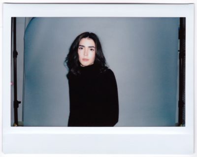 Une mannequin aux cheveux noirs mi-longs, portant un col roulé noir, se tient debout sur un fond gris. L'image, prise par le photographe parisien Thomas Duclert, est dotée d'une bordure de style Polaroïd, ce qui lui donne un aspect vintage. La mannequin a une expression neutre et est légèrement tournée vers la droite.
