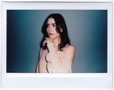 Une mannequin aux cheveux longs et foncés regarde sur le côté, posant devant un fond bleu-gris uni. Elle porte un pull rose clair à épaules dénudées dont une manche tombe sur son épaule tandis que sa main repose doucement sur sa joue. Cette photo de style Polaroïd de Thomas Duclert capture magnifiquement l'élégance parisienne.