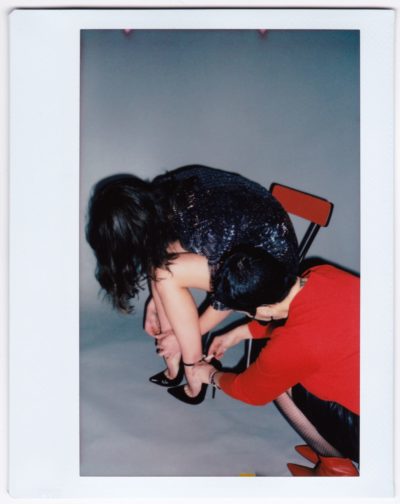 Dans une scène capturée par le photographe Thomas Duclert, une mannequin vêtue d'une robe scintillante est assise sur une chaise rouge, ses chaussures à talons hauts étant ajustées par une autre mannequin portant un haut rouge. Le mur de fond de couleur claire complète parfaitement ce moment de mode élégant.