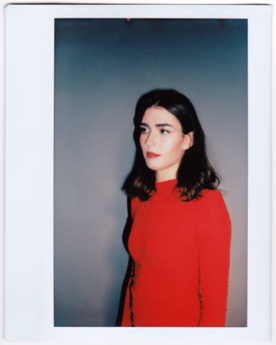 Une mannequin aux cheveux noirs mi-longs, vêtue d'une robe rouge à manches longues, se tient debout sur un fond gris uni. Elle regarde vers la gauche d'un air contemplatif. La photo, qui rappelle un Polaroïd de Thomas Duclert, a une bordure blanche.