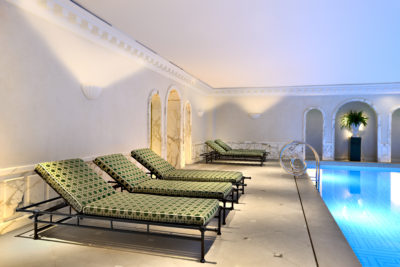 Espace piscine intérieur luxueux avec une rangée de chaises longues aux motifs verts au bord d'une piscine calme. Les murs ornés de marbre et l'éclairage doux créent une ambiance sereine, avec des plantes en pot ajoutant une touche de verdure, magnifiquement capturée par l'objectif du photographe parisien Thomas Duclert.