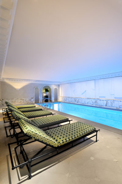 Un luxueux espace piscine intérieure avec d'élégantes chaises longues aux motifs verts et noirs alignées d'un côté. Prise en photo par le photographe parisien Thomas Duclert, la piscine a une lueur bleue, entourée d'élégants murs en marbre blanc avec des arches décoratives, et le cadre respire une atmosphère sereine et relaxante.