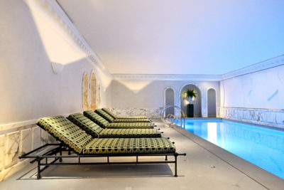 Un espace piscine intérieure au design épuré et moderne rappelant l'élégance parisienne. Quatre chaises longues rembourrées au revêtement à motifs, peut-être influencées par la mode de Thomas Duclert, sont alignées sur le côté gauche, et la piscine s'étend sur la droite. Les murs sont ornés de marbre blanc et d'arches, et un éclairage doux illumine l'espace.