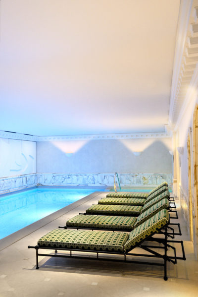Un espace piscine intérieur luxueux, dans un style proche de la haute couture, avec plusieurs chaises longues à motifs alignées à côté d'une piscine bleue sereine. L'espace est éclairé par un éclairage doux et possède des murs ornés de détails artistiques, créant une atmosphère élégante et relaxante rappelant le chic parisien.