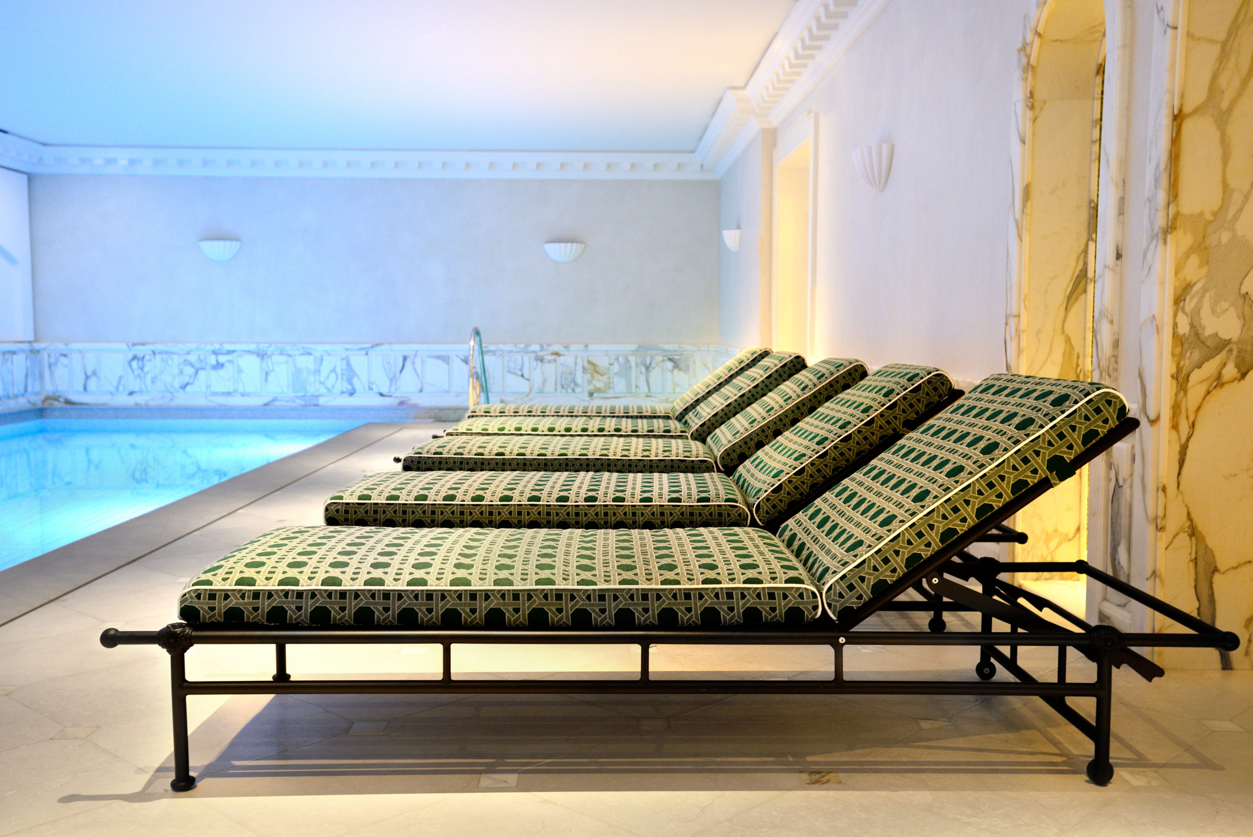 Une rangée de cinq chaises longues aux motifs verts et noirs est disposée au bord d'une piscine intérieure. L'espace piscine est doté d'un éclairage élégant, d'accents en marbre et d'une ambiance sereine rappelant le luxe parisien. Les murs, inspirés de la mode de Thomas Duclert, présentent des détails complexes qui renforcent l'atmosphère sophistiquée.