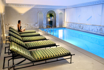 Une mannequin en maillot de bain noir, incarnant la mode parisienne, est assise au bord d'une rangée de chaises longues à motifs, à côté d'une piscine intérieure aux eaux turquoise. L'espace piscine, conçu par Thomas Duclert, comprend des murs en marbre, des portes cintrées et une plante en pot, créant une atmosphère élégante et tranquille.