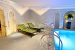 Une luxueuse salle de spa intérieure à Paris, avec deux chaises longues à motifs verts et des cadres noirs, placées à côté d'une piscine bleue illuminée. La pièce est décorée de marbre élégant sur les murs et les arcades environnantes. Conçue par Thomas Duclert, une plante en pot se trouve dans le coin sur un piédestal.