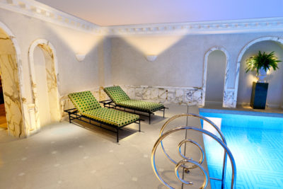 Une luxueuse salle de spa intérieure à Paris, avec deux chaises longues à motifs verts et des cadres noirs, placées à côté d'une piscine bleue illuminée. La pièce est décorée de marbre élégant sur les murs et les arcades environnantes. Conçue par Thomas Duclert, une plante en pot se trouve dans le coin sur un piédestal.