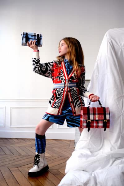 Une jeune fille se tient debout, confiante, vêtue d'une tenue élégante composée d'un pull à rayures zébrées rouges et noires, d'une jupe à motifs bleus et de bottes blanches. Elle tient une pochette à carreaux bleus dans une main et un sac à main à carreaux rouges dans l'autre, comme si elle posait pour un photographe parisien sur un fond neutre.