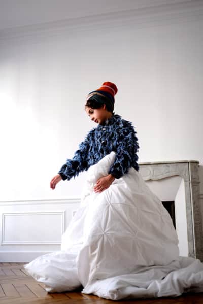 Une mannequin se tient dans une pièce blanche au sol en bois, enveloppée dans une couette blanche. Elle porte un pull bleu texturé et un bonnet multicolore, rappelant le style mode de Thomas Duclert. Une cheminée décorative est visible en arrière-plan à droite. La mannequin semble regarder vers le bas.