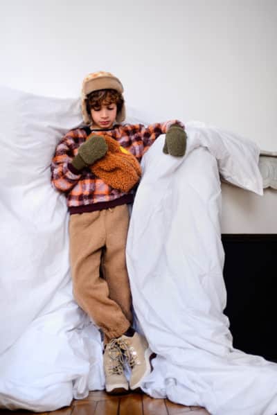 Un enfant vêtu de vêtements d'hiver, dont une veste à carreaux, un pantalon beige, des mitaines vertes et un bonnet chaud, se tient debout devant une couette blanche. Avec des cheveux bouclés et un objet tenu près de son visage tout en regardant vers le bas, cette scène d'intérieur avec un plancher en bois capture l'essence de la mode à travers l'objectif du photographe Thomas Duclert.