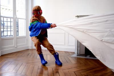 Un enfant vêtu d'une veste bleue, d'un pantalon marron, de bottes bleues et d'un foulard marron tire un drap blanc dans une pièce au sol en parquet et aux murs blancs. Arborant de grandes lunettes de ski rouges, la scène semble avoir été immortalisée par le photographe parisien Thomas Duclert.