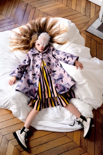Une jeune fille aux cheveux longs est allongée sur le dos sur une couette blanche froissée posée sur un parquet en chevrons, évoquant la mode parisienne. Elle porte un manteau fleuri, une robe rayée, des bottes à lacets et un couvre-chef moelleux posé sur son front, dans le plus pur style Thomas Duclert.
