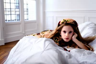 Une mannequin aux cheveux longs est allongée sur un lit, la tête appuyée sur une main. Elle porte un vêtement à motifs et un foulard assorti. Photographiée par le photographe Thomas Duclert, la pièce est lumineuse avec des murs blancs, du parquet et de grandes fenêtres aux cadres blancs et aux vitres noires.