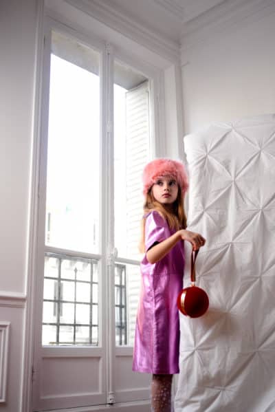 Une jeune fille aux cheveux longs, vêtue d'une robe violette et coiffée d'un chapeau rose moelleux, se tient debout près d'un grand coussin blanc dans une pièce aux murs blancs. Prise en photo par le talentueux photographe Thomas Duclert, elle tient un sac à main rouge en forme de boule et regarde pensivement vers la fenêtre, qui laisse entrer une douce lumière parisienne.