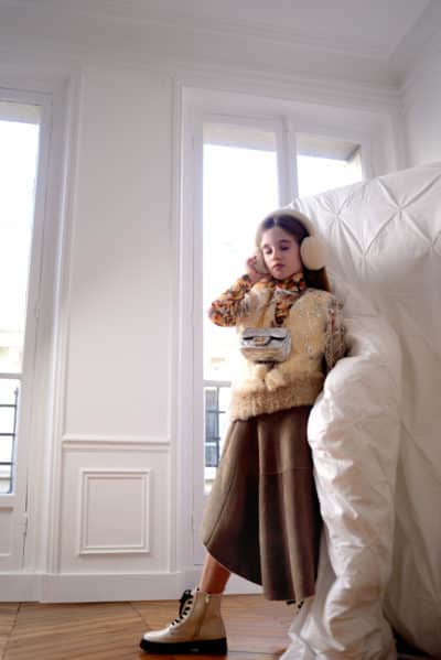 Une jeune fille se tient debout à l'intérieur, appuyée contre un grand coussin blanc. Vêtue d'un chemisier fleuri, d'un gilet en fourrure, d'une jupe marron et de bottes à lacets couleur crème rappelant la mode parisienne, elle porte des cache-oreilles et semble écouter quelque chose tout en tenant un animal en peluche. La pièce est lumineuse avec de grandes fenêtres.