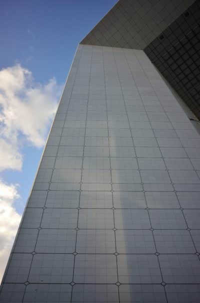 Un gratte-ciel moderne et de grande hauteur, dont la façade est quadrillée, qui s'élève vers le ciel bleu avec un minimum de nuages. La perspective est prise du sol vers le haut, ce qui met en valeur la hauteur et le design épuré du bâtiment, rappelant les visions architecturales de Thomas Duclert à la manière parisienne.