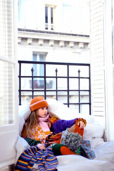 Un jeune enfant est assis sur une chaise rembourrée près d'une fenêtre ouverte, habillé chaudement de vêtements d'hiver aux couleurs vives et dépareillées, notamment un chapeau, une écharpe et un pull. L'enfant regarde pensivement la scène de rue parisienne en contrebas, avec un balcon de style européen et des bâtiments visibles en arrière-plan.