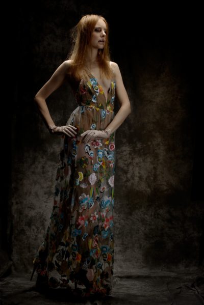 Une mannequin aux longs cheveux auburn se tient debout, confiante, une main sur la hanche. Elle porte une robe transparente sans manches ornée de broderies florales colorées. L'arrière-plan est sombre et texturé, mettant en valeur sa pose élégante et les détails complexes de sa robe, une véritable photographie de mode tout droit venue de Paris.