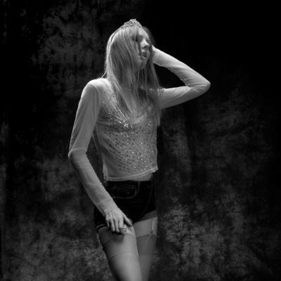 Une magnifique photo en noir et blanc montre une mannequin aux cheveux longs, portant une petite couronne, un haut transparent et scintillant, un short sombre et des jarretelles. Avec un bras levé pour toucher ses cheveux, elle prend une pose contemplative sur un fond texturé, une incarnation de la mode parisienne capturée par le photographe.
