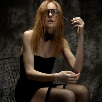 Une mannequin aux longs cheveux roux et aux lunettes est assise sur une chaise à structure métallique sur un fond sombre et texturé. Elle porte une tenue noire sans bretelles avec des bas et un collier noir, posant avec une main près du visage et l'autre bras légèrement levé, photographié par le célèbre photographe parisien Thomas Duclert.
