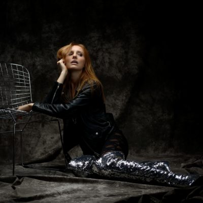 Une mannequin aux cheveux longs et roux pose de manière dramatique à genoux devant une chaise en fil de fer. Elle porte une veste en cuir noir et des bottes hautes à paillettes étincelantes. Capturée par le photographe Thomas Duclert, la toile de fond sombre et texturée ajoute une atmosphère maussade à cette scène d'inspiration parisienne.