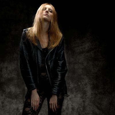 Une mannequin aux longs cheveux blonds roux se tient debout sur un fond sombre et texturé. Elle porte une veste en cuir noir et une tenue à motifs en dentelle. Capturant l'essence de la mode, le photographe Thomas Duclert cadre son expression sérieuse tandis qu'elle incline légèrement la tête en arrière, les mains posées sur les côtés.
