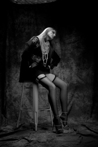 Photographie en noir et blanc d'une mannequin aux cheveux longs assise sur un grand tabouret. Elle porte une robe en dentelle, des colliers superposés et des bas porte-jarretelles montant jusqu'aux cuisses avec des talons hauts. L'arrière-plan est un tissu texturé et sombre. La pose détendue de la mannequin et l'inclinaison de la tête évoquent la mode parisienne, capturée par un photographe habile.