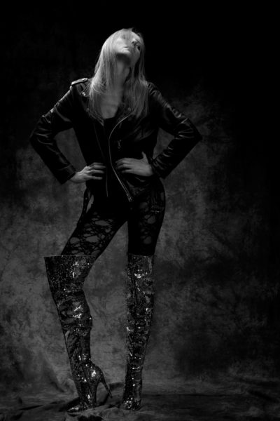 Une photo en noir et blanc d'une mannequin qui prend une pose confiante. Elle porte une veste en cuir, des leggings à motifs et des bottes à paillettes qui lui arrivent aux genoux. Ses mains sont sur ses hanches et sa tête est légèrement penchée en arrière, ses cheveux longs couvrant partiellement son visage.