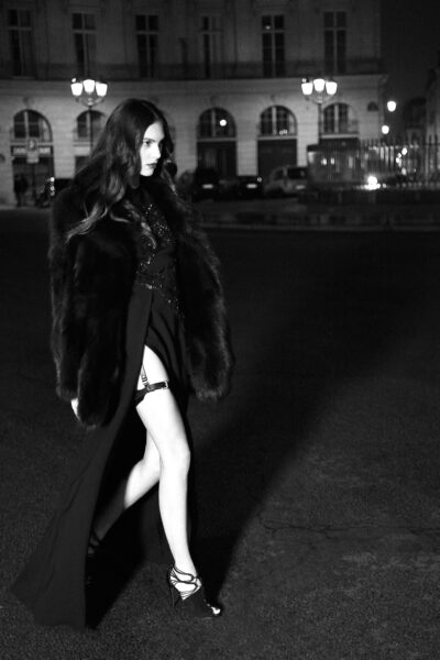 Photographie en noir et blanc d'une mannequin marchant avec assurance dans une rue de Paris la nuit . Elle porte une longue robe sombre fendue, des talons hauts et un manteau de fourrure. Des lampadaires et un immeuble aux fenêtres cintrées sont visibles à l'arrière-plan.