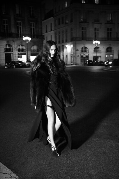 Cette mannequin vêtue d'une élégante robe de soirée noire et d'un manteau de fourrure marche avec assurance dans la nuit, dans une rue déserte de Paris. Les lampadaires l'illuminent tandis qu'elle avance, projetant une ombre douce sur le sol.