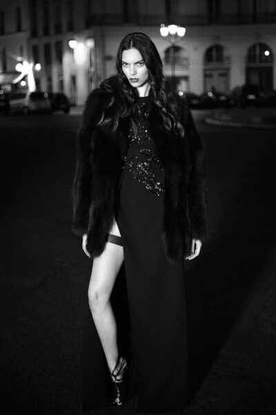 Cette mannequin en robe de soirée noire et manteau de fourrure pose avec assurance dans un décor urbain nocturne. La rue et les immeubles sont doucement éclairés en arrière-plan, créant une atmosphère dramatique et élégante.