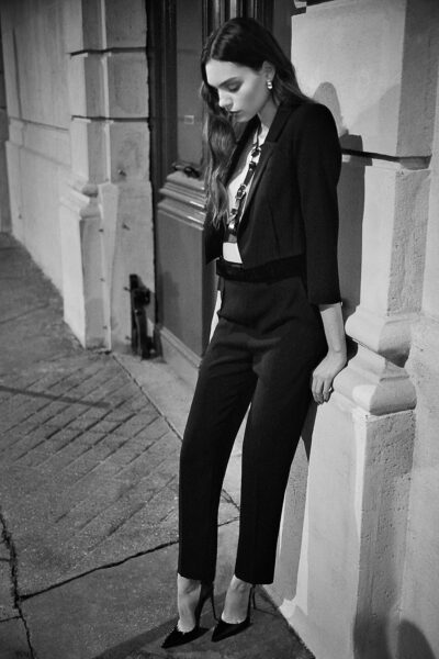 Cette mannequin en tailleur noir élégant et talons hauts s'appuie contre un immeuble dans une rue faiblement éclairée de Paris. Elle a les cheveux longs et regarde vers le bas, créant une ambiance contemplative sur cette photo en noir et blanc.