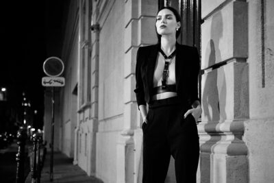 Cette mannequin d'une élégante tenue noire se tient debout, la nuit, contre un mur de pierre dans une rue de Paris faiblement éclairée. Elle a les mains dans les poches et regarde au loin. Des ombres se projettent sur le mur derrière elle.
