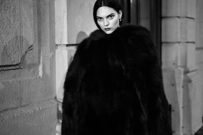 Cette mannequin en noir et blanc d'une personne portant un épais et luxueux manteau de fourrure, debout sur un trottoir de Paris. Elle a les cheveux noirs et une expression sérieuse, avec un immeuble en pierre en arrière-plan.